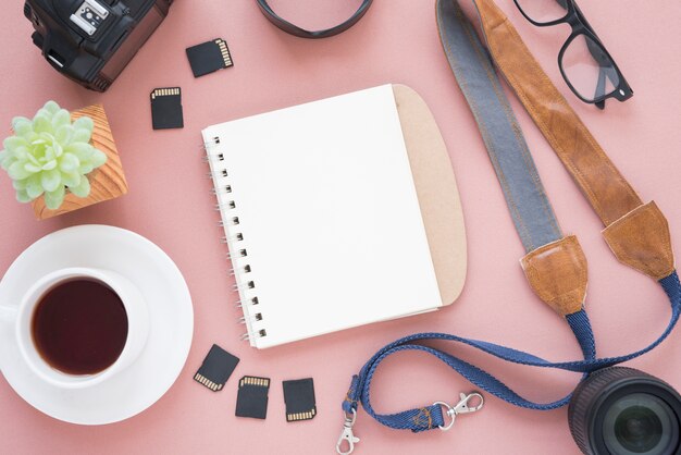 Cup of a tea; blank spiral notepad; succulent plant; camera; memory cards; camera lens; spectacle and belt over pink background