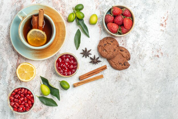 a cup of tea bowls of berries citrus fruits star anise and cinnamon sticks chocolate cookies next to the cup of tea with cinnamon on the table