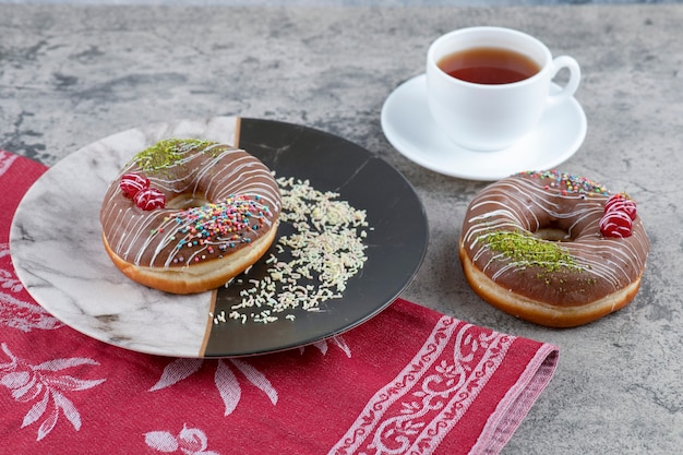Cup of tea and chocolate donuts with berry and sprinkles on marble surface.