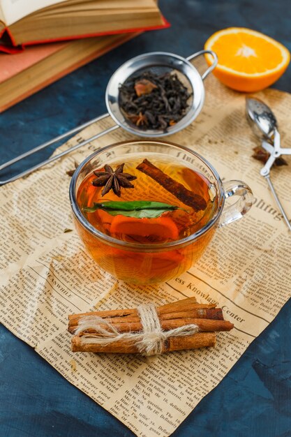 Cup of tea, cinnamon and tea strainers with newspaper, orange and a book on blue marble surface