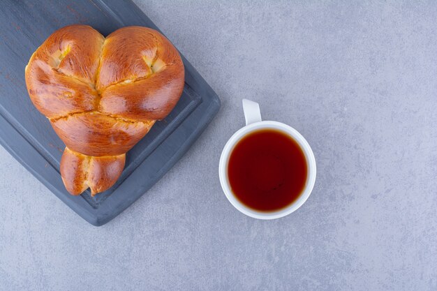 A cup of tea and a heart-shaped sweet bun on a navy board on marble surface