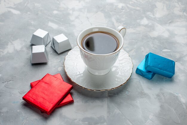 ガラスプレート上の白いカップの中に熱いお茶のカップとライトデスクの上の銀色のパッケージチョコレートキャンディー、お茶を飲む甘いクッキーティータイム