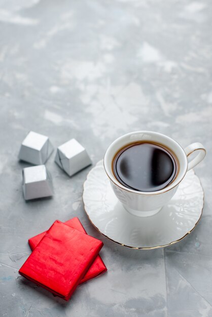 ガラスプレート上の白いカップの中に熱いお茶のカップとシルバーパッケージチョコレートキャンディーライトフロアティードリンク甘いチョコレートティータイム