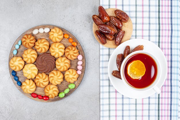 A cup of tea on a towel with dates and a wooden tray of arranged cookies and candy on marble background. High quality photo