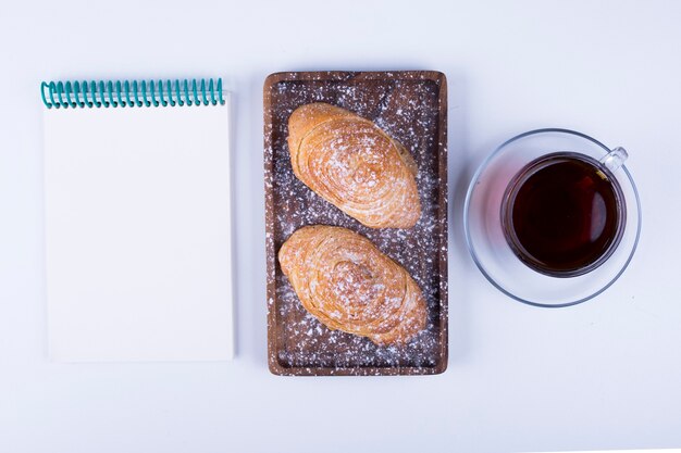 A cup of tea with badamburas, a blank notebook aside in plate on white wooden background