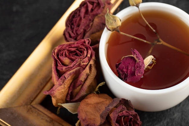 黒いテーブルの上に乾燥したバラとフレームのお茶のカップ。