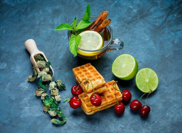 Cup of tea with mint, cinnamon, dried herbs, cherry, lime on grungy blue surface, flat lay.