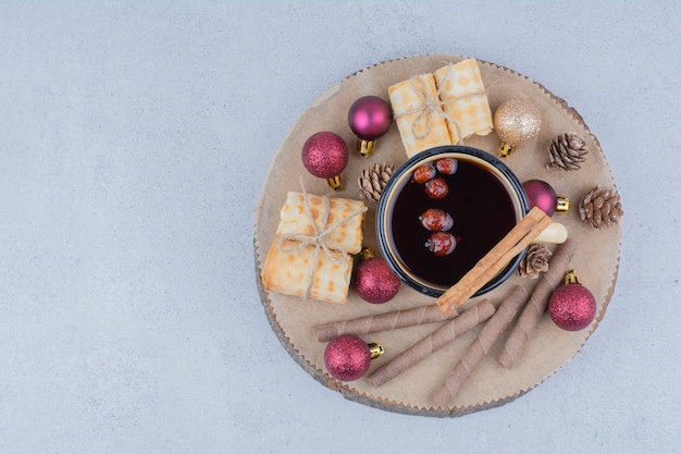 Чашка чая с шиповником, печеньем и шарами на деревянной доске.