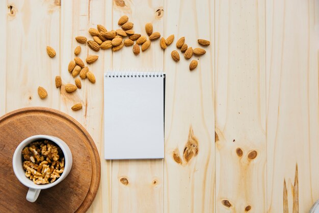 Cup of walnut near blank notepad and almonds on wooden backdrop