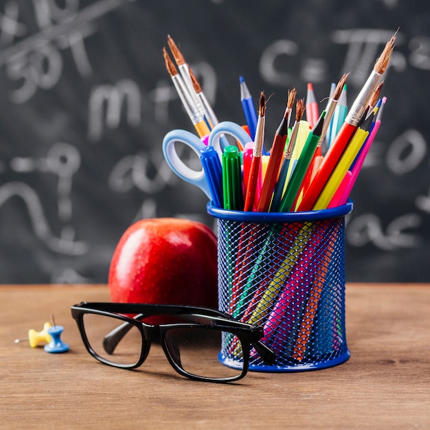 Cup with colourful stationery near glasses and apple on table