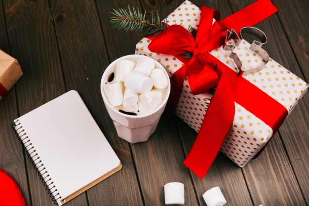 Cup with hot chocolate and marshmallows stands on the floor before present box and empty notebook