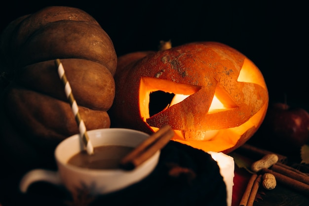 Cup with hot chocolate stands before shiny scarry halloween pumpkin