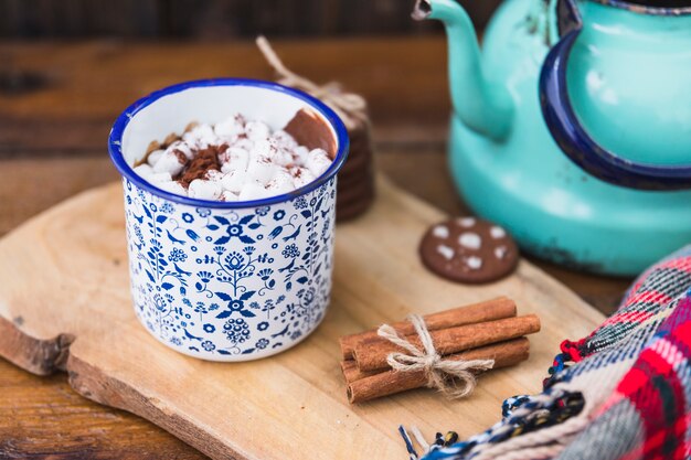 Cup with marshmallow near biscuits, cinnamon and kettle