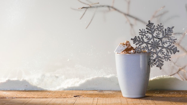 Cup with toy snowflake on wood table near bank of snow