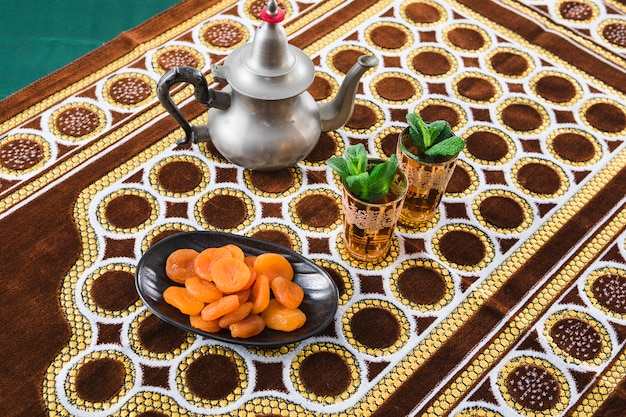 Cups of drink near teapot and dried apricots on mat 