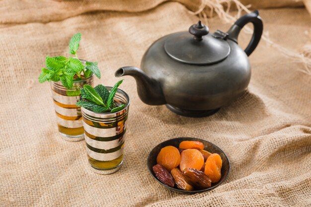 Cups near retro teapot and dried fruits near mat on wrinkled burlap