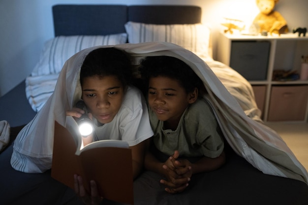 Curious african american kids lying together on bed and reading book with flashlight at night