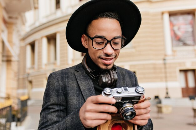 카메라를보고 모자에 호기심 아프리카 남자. 아름 다운 건물 근처에 서 흑인 남성 사진 작가의 야외 초상화.