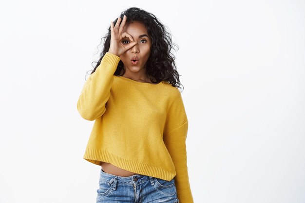 Curious amused young cute africanamerican girl in yellow sweater folding lips gasping astonished say wow hold okay circle fingers over eye and gazing through checkout interesting offer