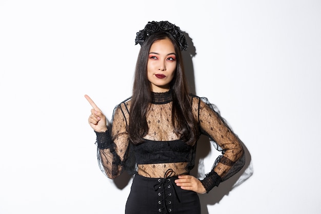 Любопытная привлекательная азиатская женщина в костюме ведьмы, указывая пальцем в верхнем левом углу, заинтересованная в промо на хэллоуин, стоит над белой стеной и думает