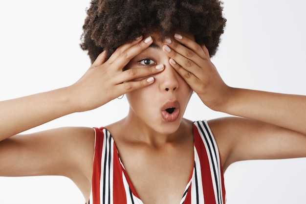 Free photo curious attractive dark-skinned female model with afro hairstyle in striped clothes covering eyes with palms and peeking through fingers with interest folding lips, being intrigued