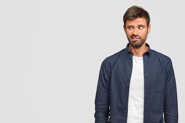 Free photo curious bearded young european male looks nervously, purses lips and gazes suspiciously aside, dressed in casual clothes, stands alone against white wall with copyspace