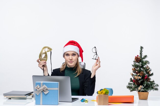 Curious beautiful woman with santa claus hat eyeglasses and mask sitting at a table with a Xmas tree and a gift on it in the office on white background