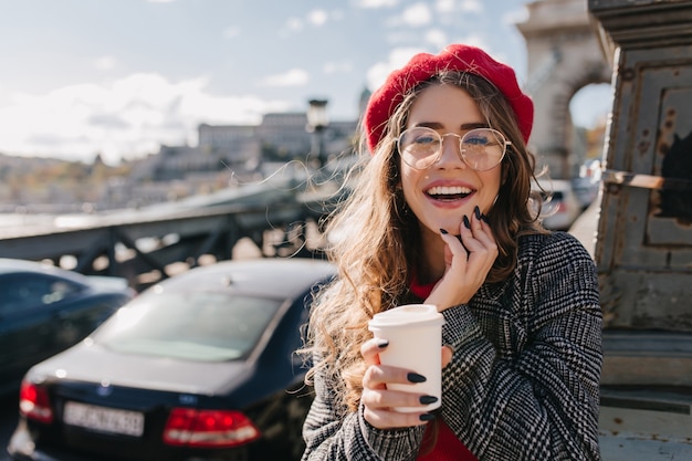 Curiosa ragazza bionda in elegante berretto rosso in posa con il sorriso su sfocatura dello sfondo in una mattina ventosa