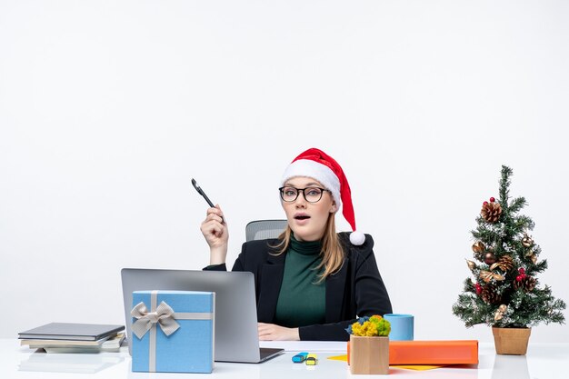Curious blonde woman with a santa claus hat sitting at a table with a Christmas tree and a gift on it focused on something on white background