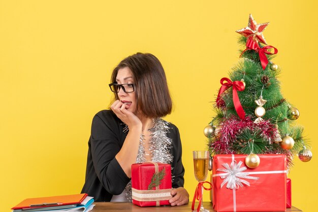 그녀의 선물을 내려다보고 사무실에서 그것에 크리스마스 트리와 함께 테이블에 앉아 안경 정장에 호기심 비즈니스 아가씨