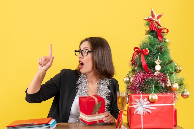 Curious business lady in suit with glasses holding her gift pointing up and sitting at a table with a xsmas tree on it in the office