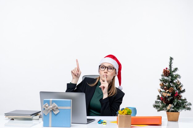 白い背景の上のオフィスでクリスマスツリーとその上に贈り物とテーブルに座っているサンタクロースの帽子を持つ好奇心旺盛なビジネス女性