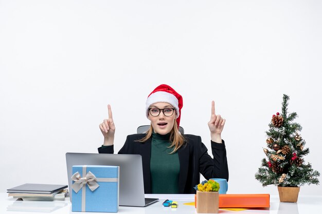 Curious business woman with santa claus hat sitting at a table with a Xmas tree and a gift on it pointing above on white background