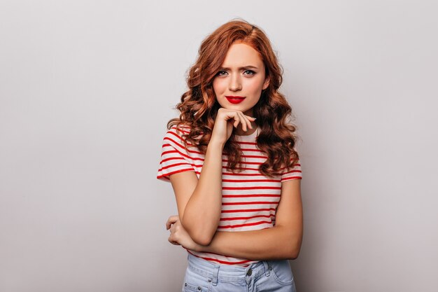 Curious caucasian girl with long wavy hair posing  Indoor photo of wonderful ginger woman isolated on light wall.