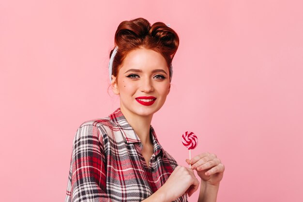 Curious caucasian woman with lollipop looking at camera. Front view of cute pinup girl with candy isolated on pink space.