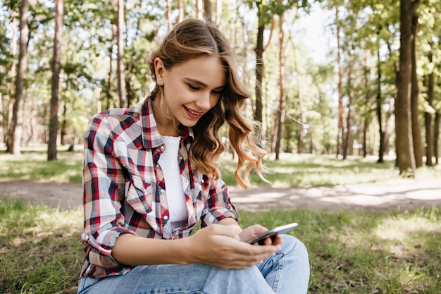 Любопытная кудрявая девушка отправляет текстовое сообщение, сидя на траве. Наружная фотография великолепной стильной дамы, отдыхающей в лесу.