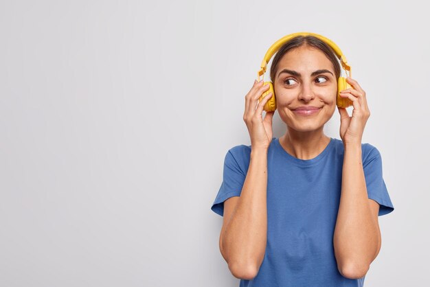 Curious dark haired woman keeps hands on yellow wireless headphones looks away dressed in casual blue t shirt enjoys favorite playlist isolated over white background copy space for your text