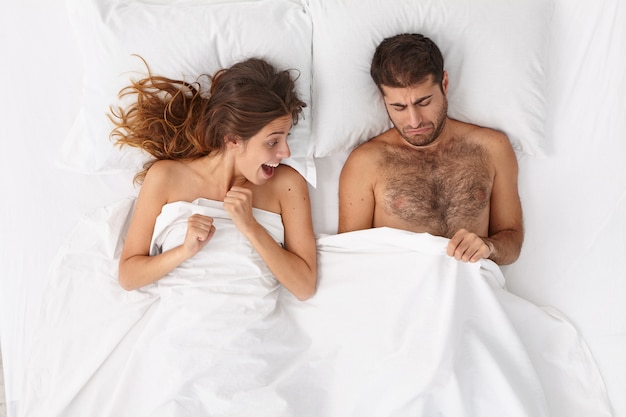 호기심이 흥분한 여자가 함께 침대에 누워있는 동안 남자의 생식기를 바라본다. 불쾌한 남자는 성기의 흰 담요 아래를보고 성기능 장애를 앓고 있습니다. 성 문제, 결혼, 관계 개념