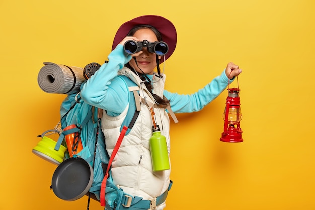 Curious female backpacker explores tourist destination, uses binoculars, dressed in active wear, holds kerosene lamp carries travelling items with rucksack