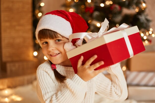 Любопытная девочка женского пола в белом свитере и шляпе санта-клауса, трясет подарочной коробкой, интересуется, что внутри, позирует в праздничной комнате с камином и рождественской елкой.