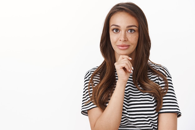 Curious good-looking cheerful female in striped t-shirt touch chin thoughtful, smiling enthusiastic and intrigued, hear amusing story, pondering as standing white wall