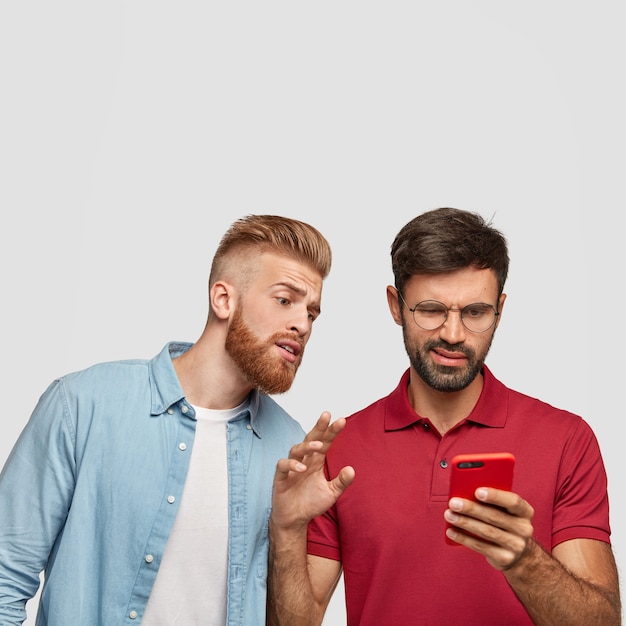 Curious guy with ginger haircut and beard peeks into smart phone of his friend