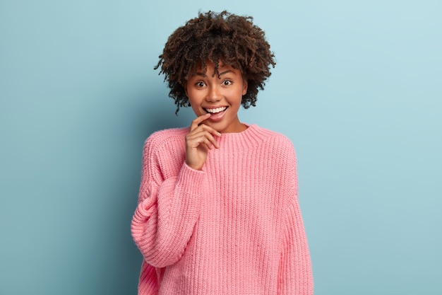 Curious happy woman keeps index finger on lips, looks with pleased facial expression, listens fine latest news from friend