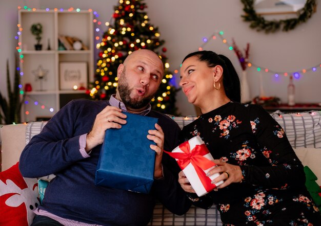 Curious husband and pleased wife at home at christmas time sitting on sofa in living room both holding gift package looking at each other