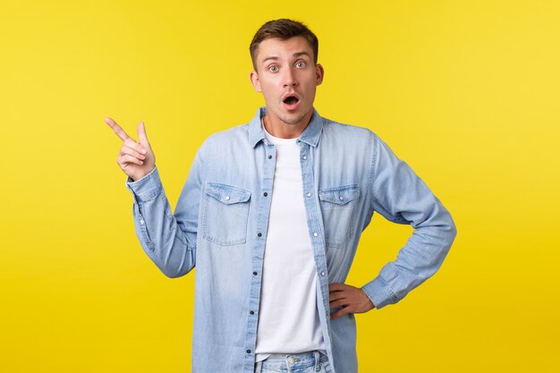 Curious and interested handsome blond guy asking question about new product or service, pointing upper left corner and looking intrigued at camera, discussing promo offer, yellow background.