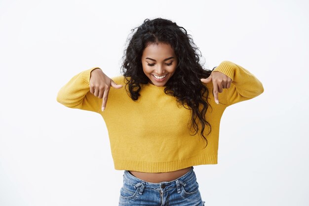 노란색 스웨터를 입은 호기심 많고 귀여운 아프리카계 미국인 소녀가 즐거운 미소로 가리키며 내려다보고, 놀라운 프로모션을 확인하고, 광고를 응시하고, 흰 벽을 바라보고 있습니다.