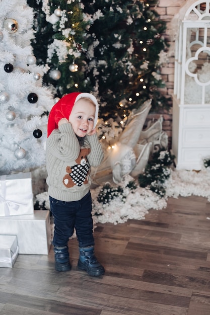 Curious little boy in Santa hat smiling and walking near Christmas tree