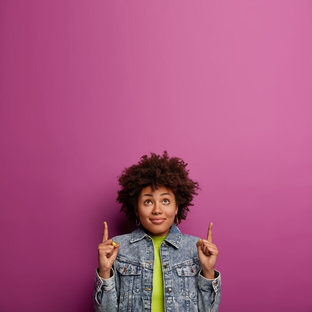 Curious lovely ethnic woman with curly hairstyle, points fore fingers upwards