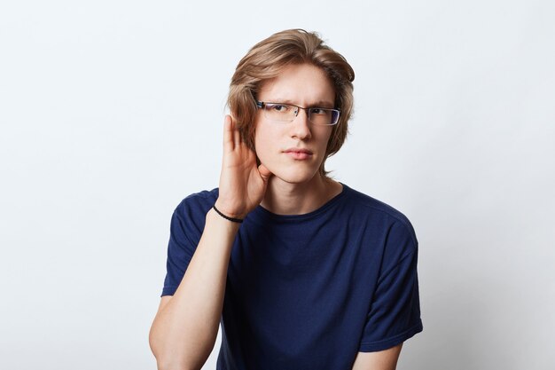 Curious male with trendy hairstyle, eavesdropping something in next room, keeping his hand near ear, trying to onhear something interesting. Stylish male manager with intriguing expression