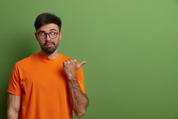 Free photo curious man looks with suspicion and interest aside, points aside on blank space, shows advertisement of product or company banner, gestures against green wall, wears glasses, t shirt.